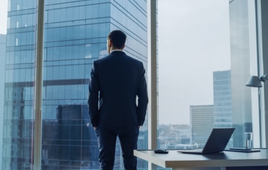 A businessman in an office looks out a large window at the cityscape