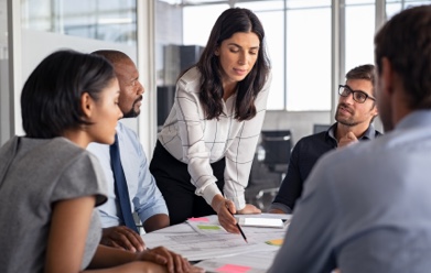 A woman points to some papers on a table while others watch her presentation.