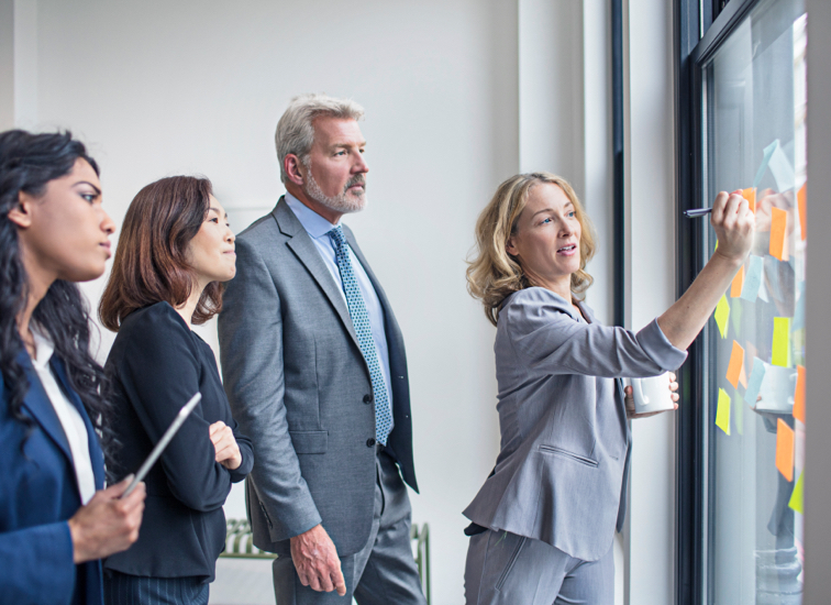 Three associates looking at Post-It Notes on a window while a fourth writes on a Post-It Note.