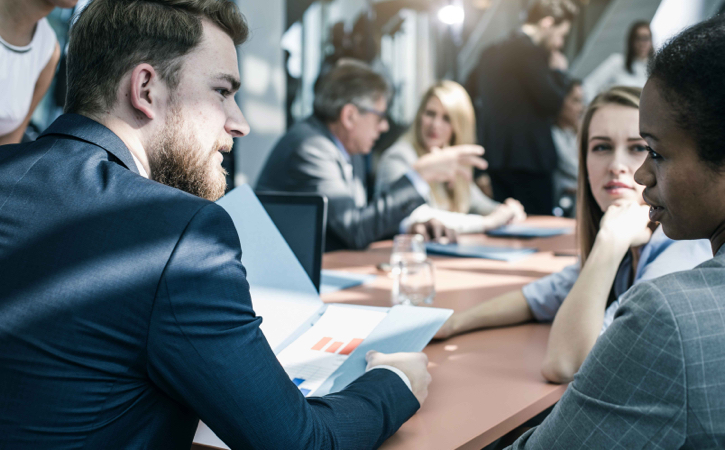 A man holds a portfolio and speaks with two women seated at a table.