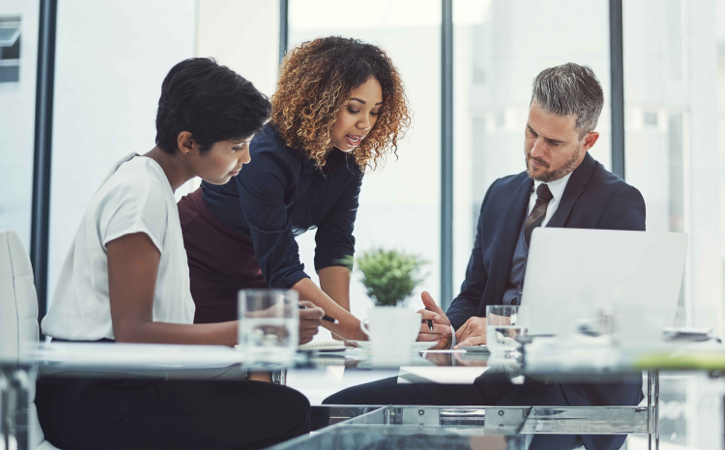A woman leans over a table and discusses business matters with a man and woman seated at a table.