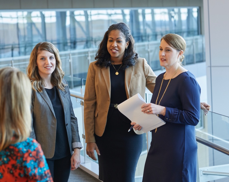 Four women standing along a glass railing inside an office building having a conversation.